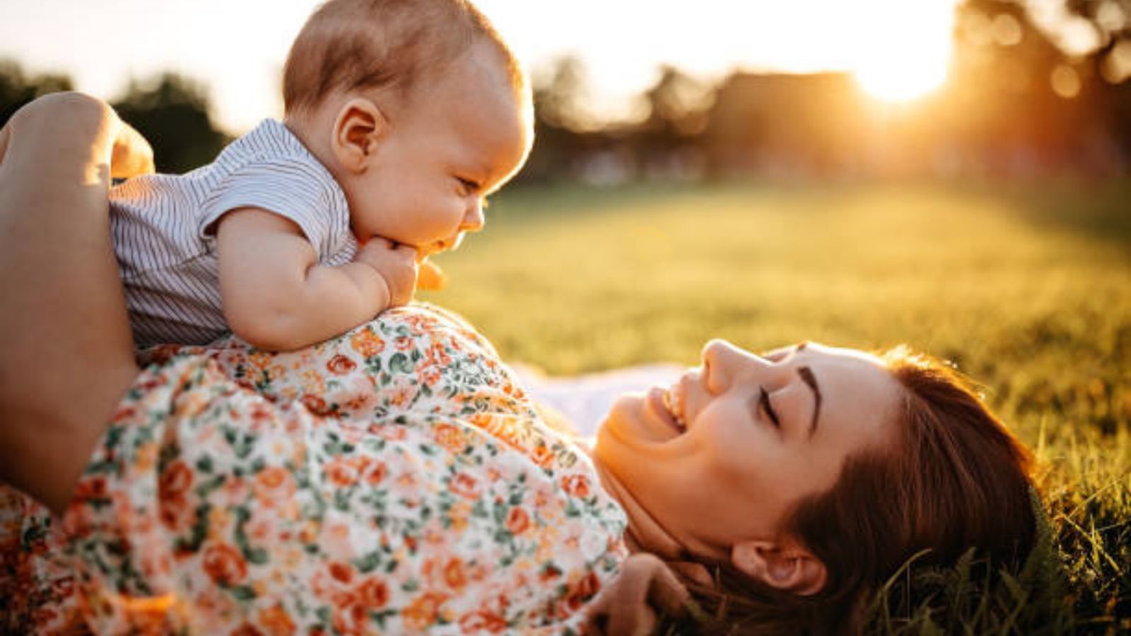 A smiling mom lying in the grass at sunset, her baby on her chest