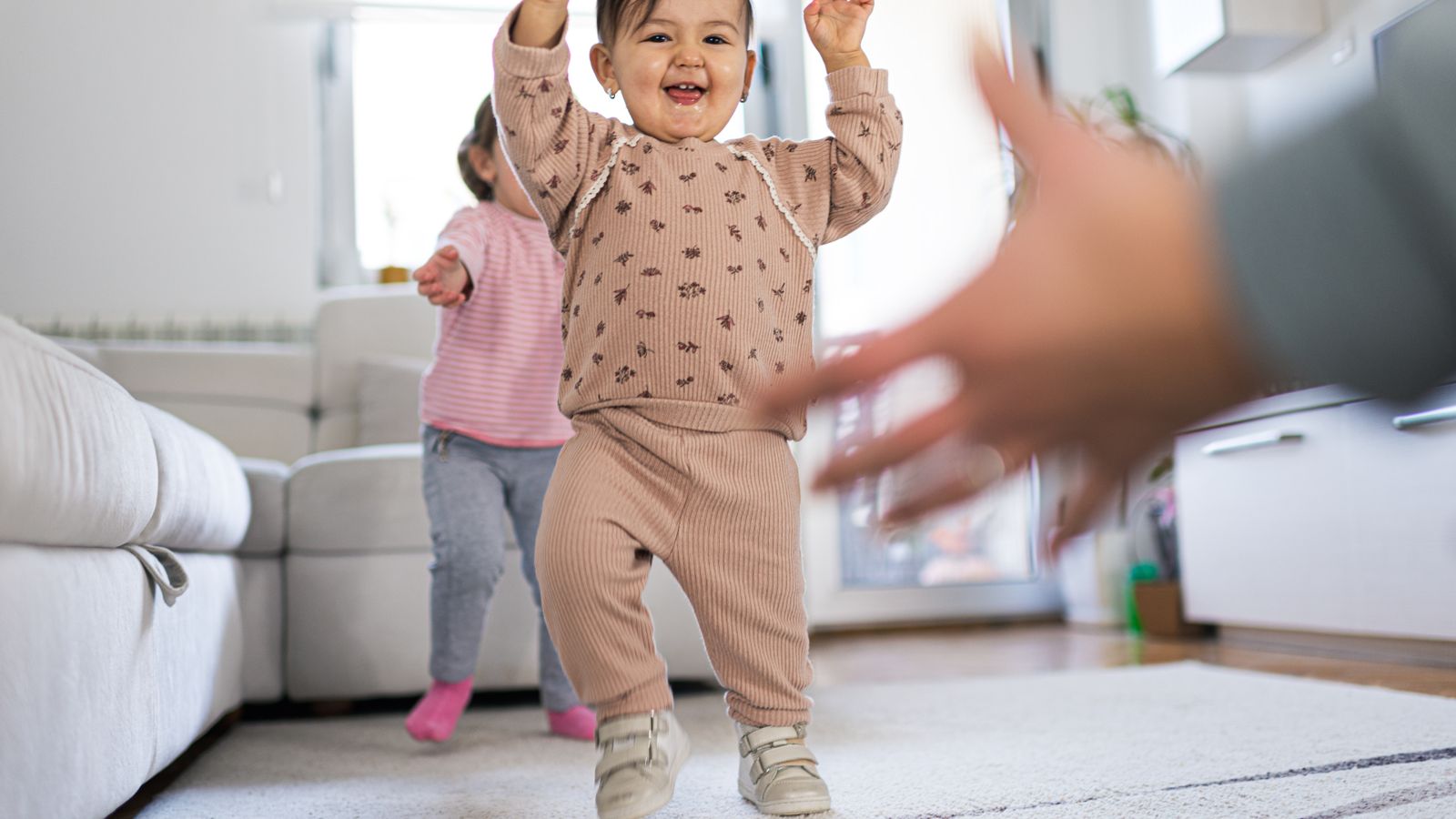 A little girl walking toward open arms, her big sister behind her