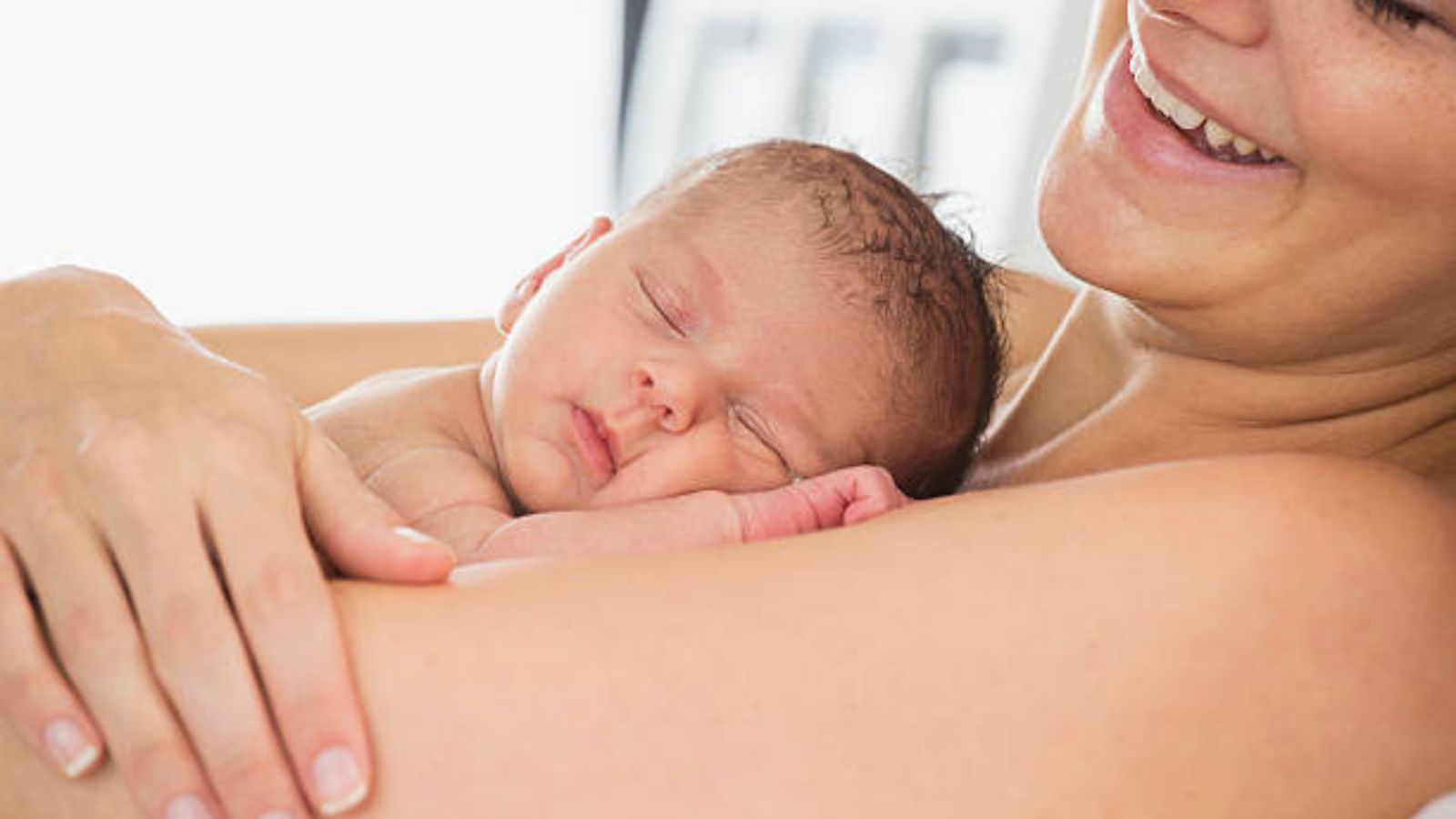 A smiling mom holding her sleeping newborn skin to skin