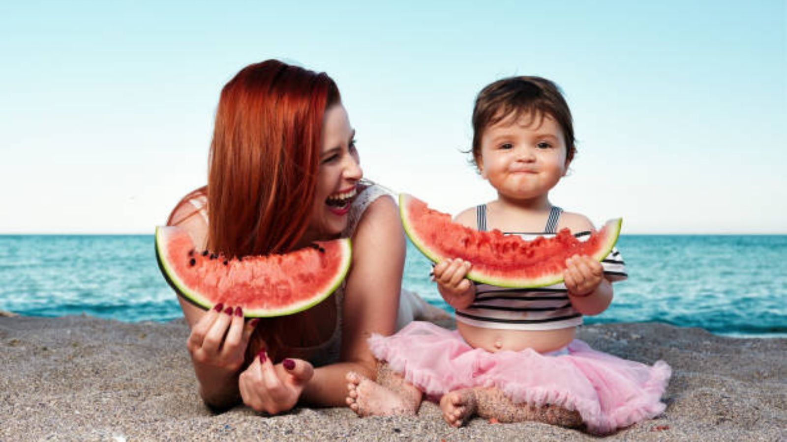 A laughing mom and her baby girl sharing slices of watermelon on the beach