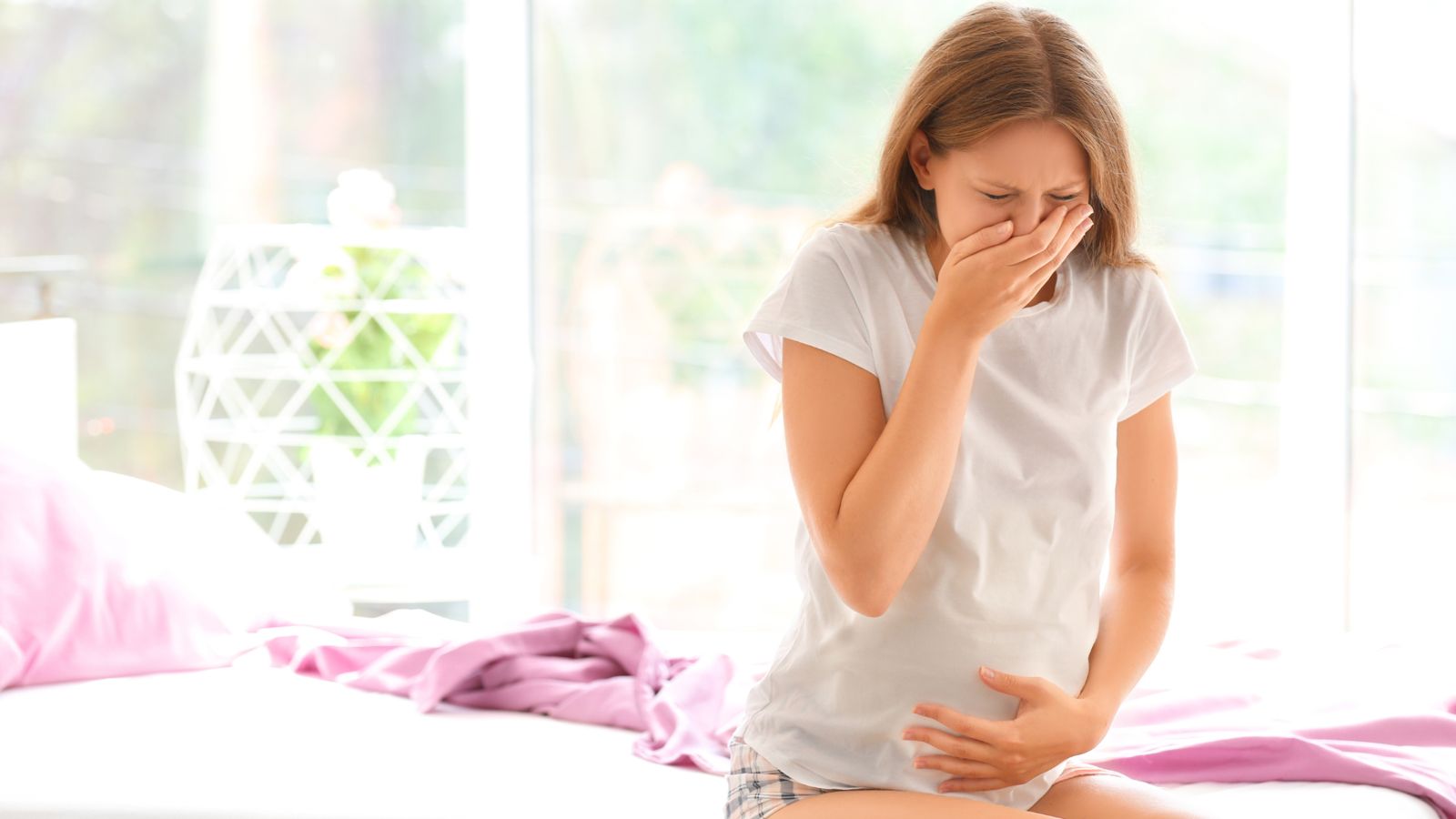 A pregnant woman feeling queasy, hand to her mouth, on the edge of her bed