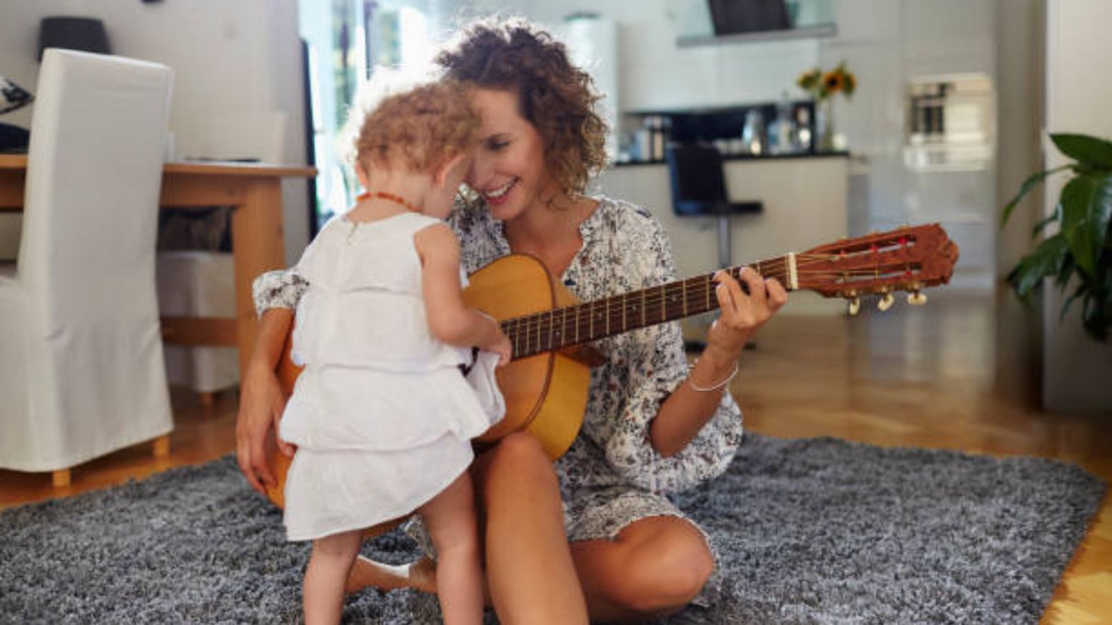 A smiling mom playing guitar on the rug for her curly-haired little girl