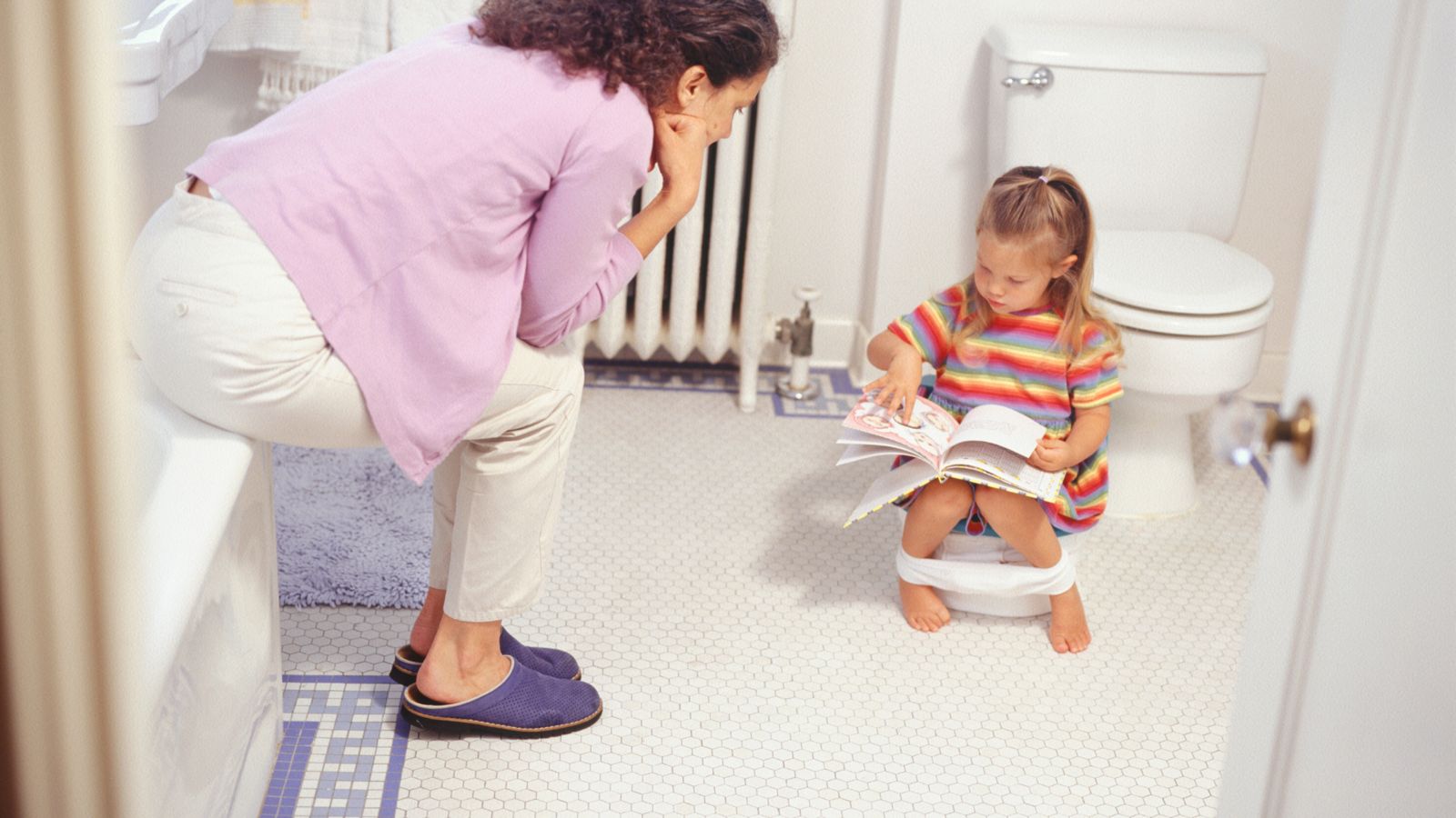A mom sitting with her daughter as she reads a book on the potty