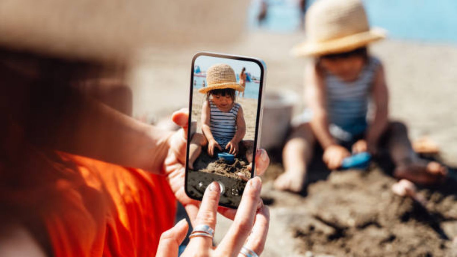 A parent taking a phone photo of a toddler playing in the sand