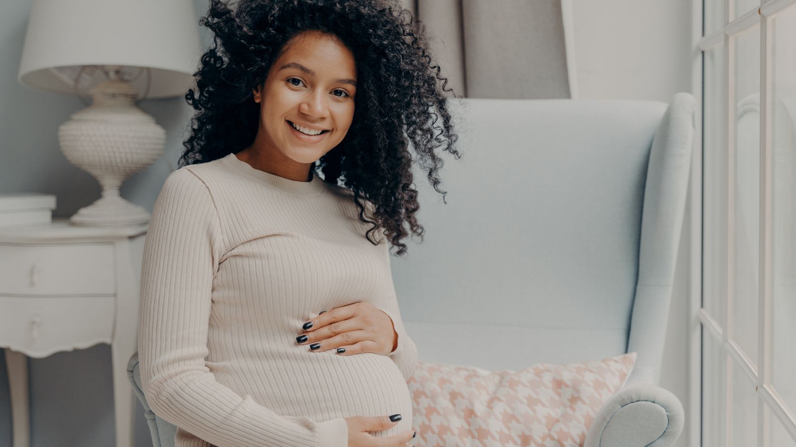 A smiling pregnant mother in a cozy nursery chair, hands on her bump