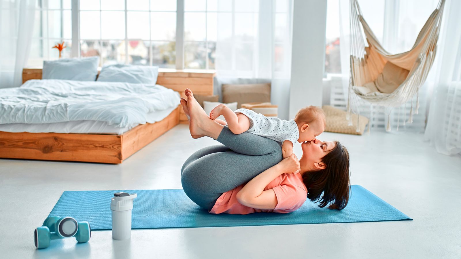 A mother on a yoga mat at home, kissing the baby balanced on her shins
