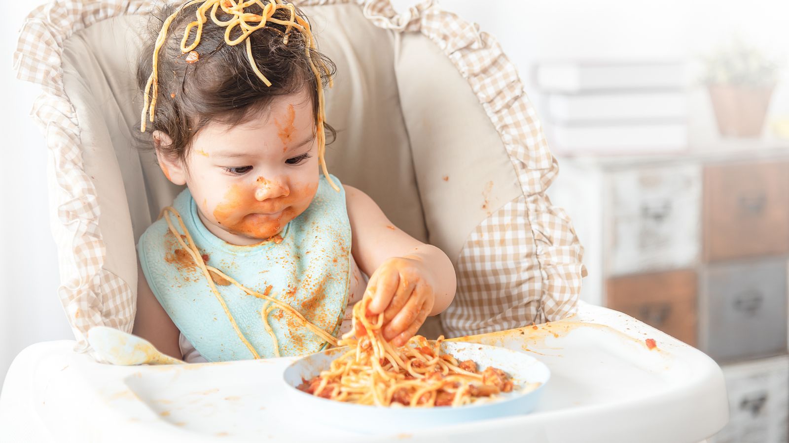 A baby girl in a high chair with spaghetti on her head, grinning into her bowl